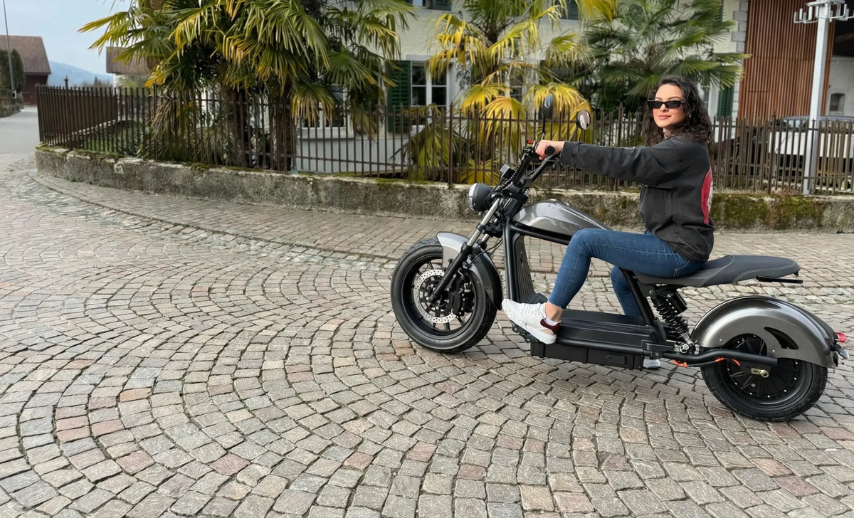 Frau mit Sonnenbrille auf einem grauen Elektro-Chopper auf Kopfsteinpflaster